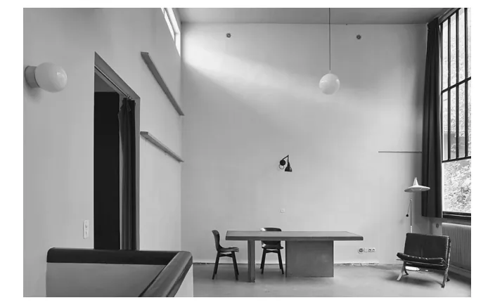 Zwart-witfoto van een hoog atelier met een lange tafel, stoelen, een bank en hangende lampen, met grote ramen aan de rechterkant.