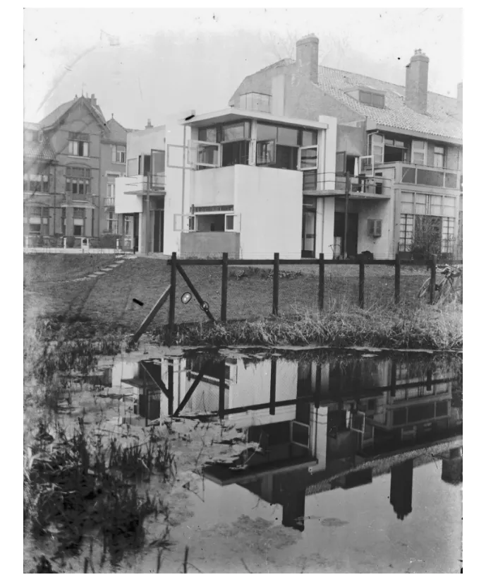 Zwart-witfoto van het Rietveld-Schröderhuis uit de tijd van oplevering, met witte gepleisterde muren, een plat dak en een hek met een weerspiegeling in het water op de voorgrond.