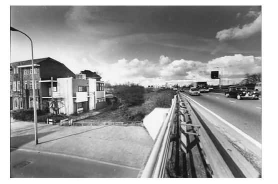 Zwart-witfoto van een brede weg met een geleiderail en een lantaarnpaal, met op de achtergrond het Rietveld-Schröderhuis en een woonblok.