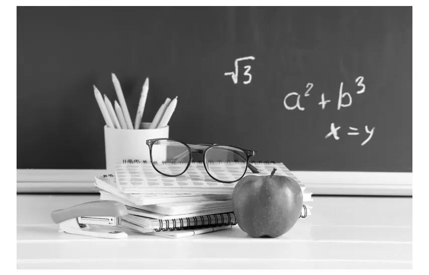 Zwart-witfoto van een stapel schriften met een bril, een appel, een nietmachine en een beker potloden voor een schoolbord met wiskundige formules.