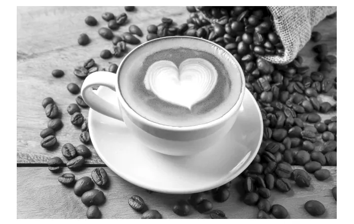 Zwart-witfoto van een kop koffie met een hartje in het melkschuim op een schotel, omringd door koffiebonen en een jute zak.