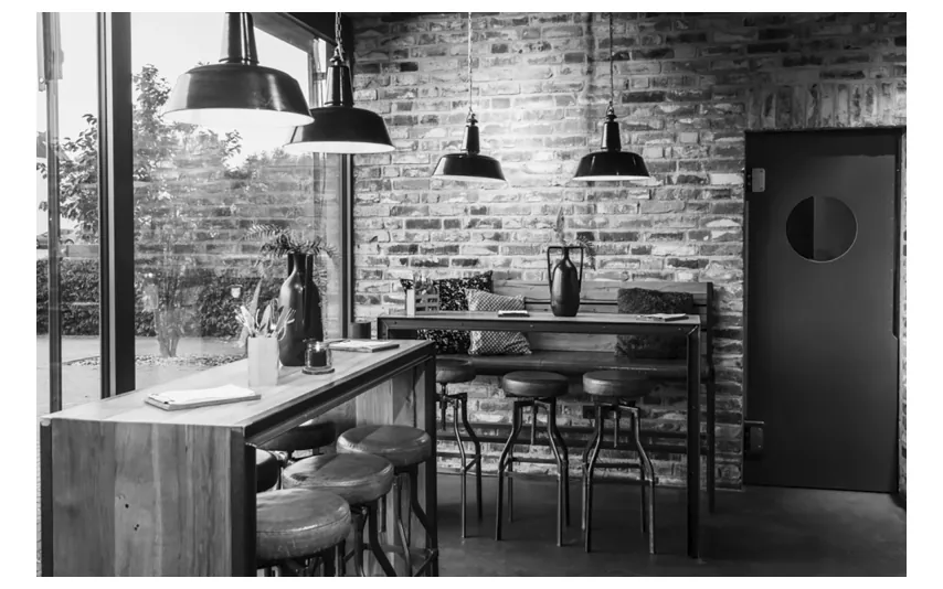 Zwart-witfoto van een horecazaak met een bakstenen muur, hanglampen, een houten bar met krukken en grote ramen naar buiten.
