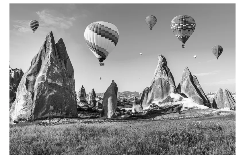 Zwart-witfoto van rotspieken (feeënschoorstenen) in Cappadocië met een grasveld op de voorgrond en heteluchtballonnen in de lucht.