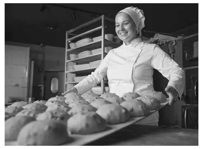 Foto van een glimlachende vrouw in bakkerskleding die een plaat met broodjes vasthoudt.