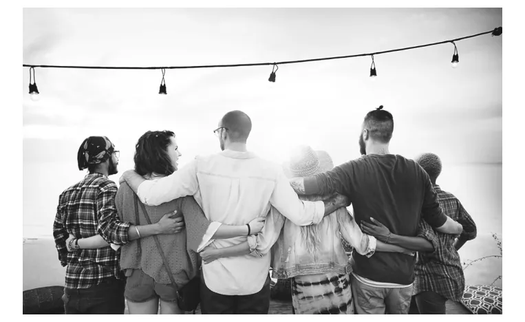 Zwart-witfoto van een groep vrienden die van achteren gezien arm in arm staan, onder een slinger met lampjes.