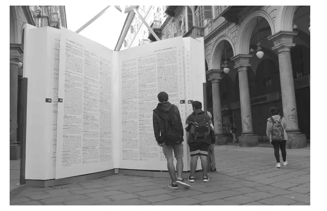 Zwart-witfoto van een groot boekmonument op een plein, met twee jongeren die ernaar kijken.
